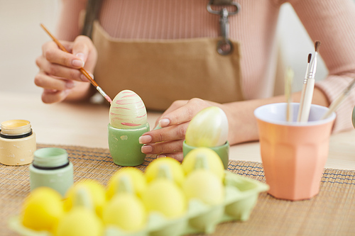 Close up of young woman painting eggs in pastel colors for Easter while sitting at table in kitchen or art studio, copy space