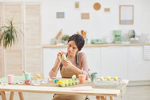 Wide angle view at elegant adult woman painting eggs in pastel colors for Easter while sitting at table in kitchen or art studio, copy space