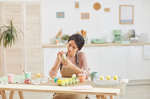 Wide angle view at elegant adult woman painting eggs in pastel colors for Easter while sitting at table in kitchen or art studio, copy space