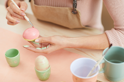Closeup of unrecognizable woman painting eggs in pastel colors for Easter while sitting at table in kitchen or art studio, copy space