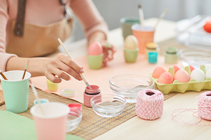 Close up of unrecognizable woman painting Easter eggs in pastel colors while sitting at table in kitchen or art studio, copy space