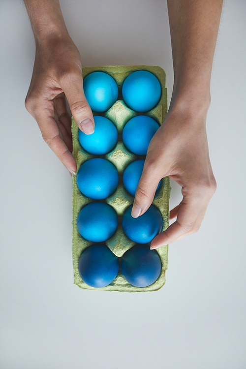 Above view background female hand arranging composition of painted blue Easter eggs in crate on white, copy space