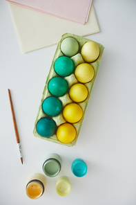 Top view of green and yellow painted Easter eggs in crate arranged in minimal composition with paint brush on white background, copy space