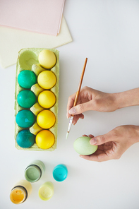 Top view of female hand painting Easter eggs in pastel green and yellow colors over white background, copy space