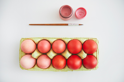 Top view of pastel pink and red Easter eggs in crate arranged in minimal composition with paint brush on white background, copy space