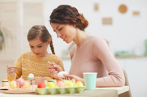 Candid portrait of mother and daughter painting Easter eggs sitting at table in cozy kitchen interior, copy space