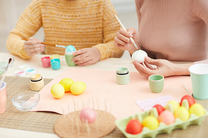 Closeup of mother and daughter painting Easter eggs sitting at table in cozy kitchen interior, copy space