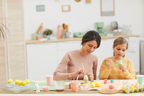 Wide angle portrait of mother and daughter painting Easter eggs pastel color sitting at table in cozy kitchen interior, copy space