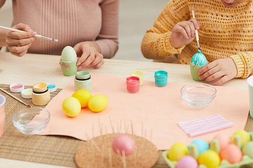 Close up of mother and daughter painting Easter eggs pastel color sitting at table in cozy kitchen interior, copy space
