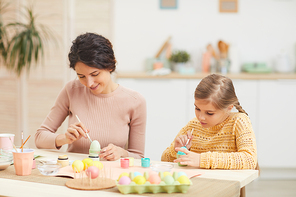 Candid portrait of mother and daughter painting Easter eggs pastel colors sitting at table in cozy kitchen interior, copy space