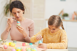 Portrait of mother and daughter painting Easter eggs together sitting at table in cozy kitchen interior, copy space