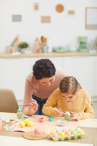 Vertical portrait of mother and daughter painting Easter eggs together sitting at table in cozy kitchen interior, copy space