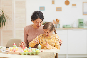 Portrait of loving mother and daughter painting Easter eggs pastel colors sitting at table in cozy kitchen interior, copy space