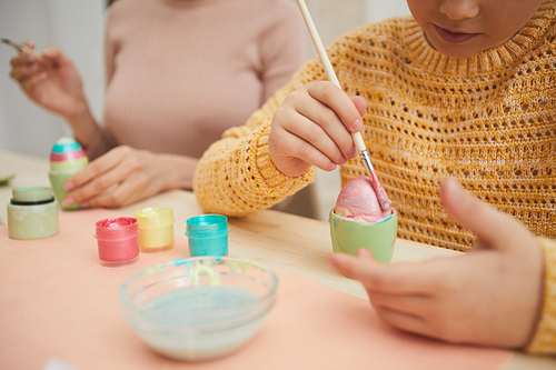 Close up of little girl painting egg pastel pink while preparing Easter decorations with loving mother at kitchen table, copy space