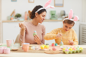 Portrait of young mother with little girl having fun while painting Easter eggs in cozy kitchen interior, both wearing bunny ears, copy space