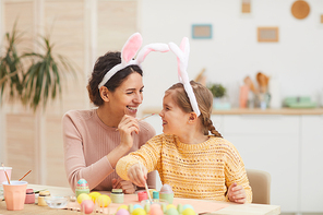 Portrait of loving mother with little girl having fun while painting Easter eggs in cozy kitchen interior, both wearing bunny ears, copy space