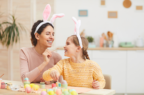 Portrait of loving mother with daughter having fun while painting Easter eggs in cozy kitchen interior, both wearing bunny ears, copy space