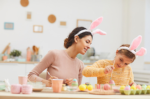Portrait of young mother with cute little girl enjoying painting Easter eggs in cozy kitchen interior, both wearing bunny ears, copy space