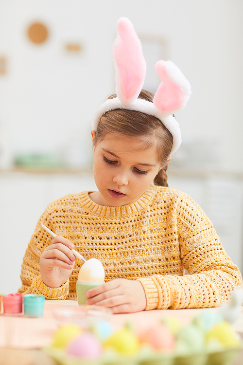 Vertical portrait of cute little girl painting Easter eggs while wearing bunny ears in cozy kitchen interior, copy space