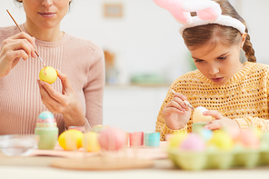 Portrait of cute little girl painting Easter eggs with mother while wearing bunny ears in cozy kitchen interior, copy space