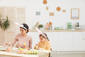 Portrait of young mother and daughter painting Easter eggs in cozy kitchen interior, both wearing bunny ears, copy space