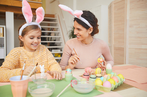 Portrait of happy young mother and daughter painting Easter eggs in cozy kitchen interior, both wearing bunny ears, copy space