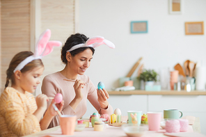 Side view portrait of happy young mother and daughter painting Easter eggs in cozy kitchen interior, both wearing bunny ears, copy space
