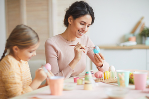 Side view portrait of happy young mother and daughter painting Easter eggs in cozy kitchen interior, copy space