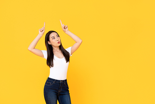 Young beautiful Asian woman pointing up with her two fingers while pouting her lips in a yellow isolated studio background