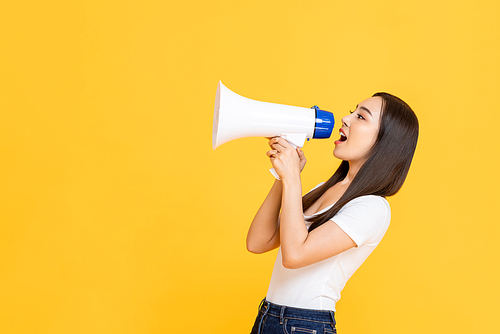 Side view portrait of young pretty Asian woman holding megaphone while shouting announcement in isolated studio yellow background