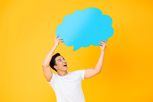 Excited smiling young handsome Asian man holding and looking up to speech bubble with empty space for text on yellow background