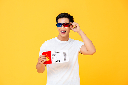 Waist up portrait of happy young Asian man wearing 3D glasses holding movie ticket isolated on yellow studio background