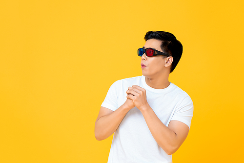 Excited young handsome Asian man wearing 3D movie glasses looking at empty space with hands together isolated on yellow background