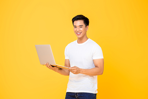 Portrait of a smiling  young handsome Asian man holding laptop computer while  in isolated studio yellow background