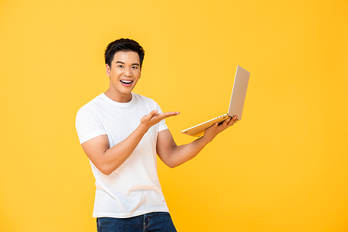 Portrait of smiling young handsome Asian man presenting laptop while smiling and looking  at camera in isolated studio yellow background