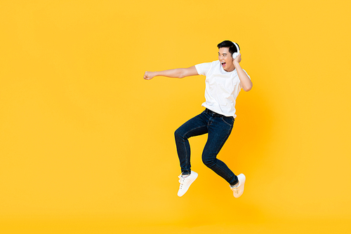 Happy handsome young Asian man wearing headphones punching and jumping in colorful yellow background