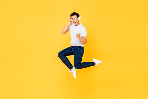 Happy young Asian man listening to music from wireless headphone and jumping isolated on yellow studio background