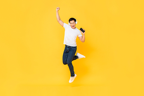 Happy young Asian man wearing headphones listening to music from mobile phone and jumping with fist raising isolated on yellow background