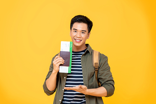 Portrait of smiling young Asian backpacker tourist man showing his passport with boarding pass in open palm gesture isolated on studio yellow background