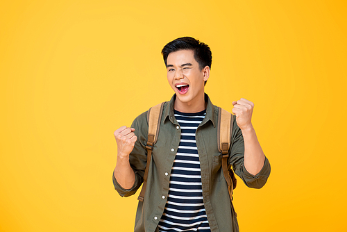 Portrait of happy excited  young Asian tourist backpacker man raising his fists doing success gesture in isolated studio yellow background