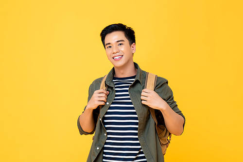 Portrait of smiling young male Asian tourist carrying backpack ready to travel in isolated studio yellow background