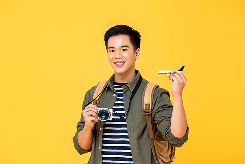 Handsome smiling Asian tourist man holding plane model and camera isolated on yellow studio background