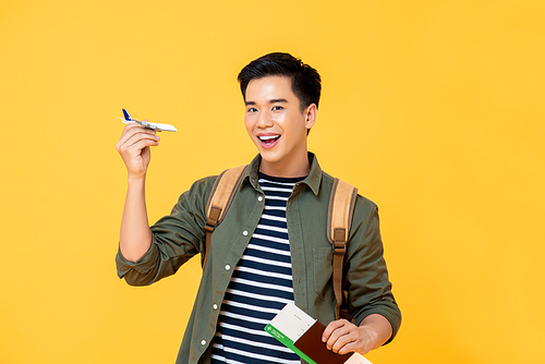 Fun travel concept portrait of smiling young male Asian tourist holding airplane toy with passport and ticket in isolated studio yellow background