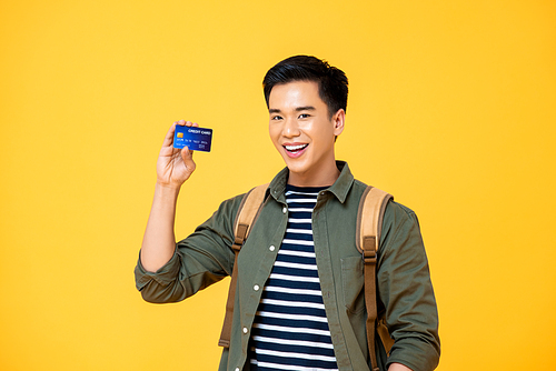 Portrait of a smiling young Asian backpacker tourist man showing credit card in isolated studio yellow background