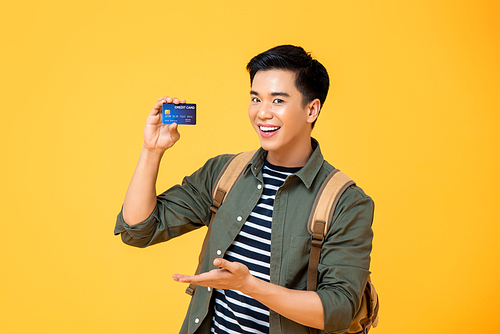 Smiling cheerful young Asian male tourist backpacker holding credit card isolated on yellow studio background