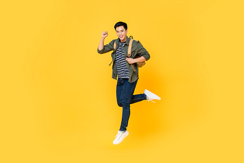 Happy smiling young Asian tourist man with backpack jumping in mid-air isolated on yellow background