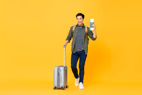 Young handsome Asian tourist man with luggage passport and boarding pass ready to go for travel on isolated yellow background