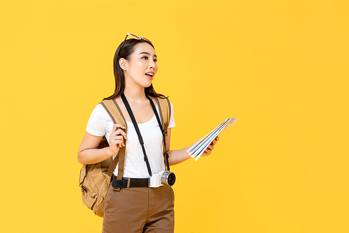 Travel concept portrait of young backpacker Asian woman tourist holding map while looking away in isolated studio yellow background