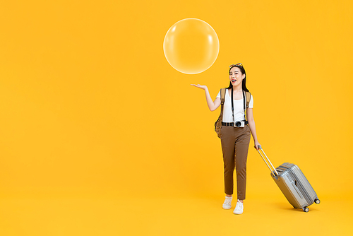 Full length portrait of  happy young Asian female tourist holding luggage with open palm showing bubble in isolated studio yellow background