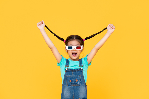 Fun Portrait of excited young girl wearing 3D cinema glasses with both arms raised pulling her hair in isolated studio yellow background